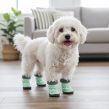 Petit chien blanc portant des bottes d'hiver imperméables vert menthe avec semelles antidérapantes, debout sur un sol en bois dans un intérieur chaleureux.