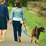 Deux personnes qui marchent sur une route avec un chien et une laisse et un distributeur et ramasseur de crottes pour chien de couleur bleu attaché sur la laisse du chien