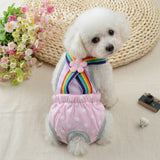 Small white-haired dog wearing a pink overall diaper with white polka dots and a multi-colored band. 